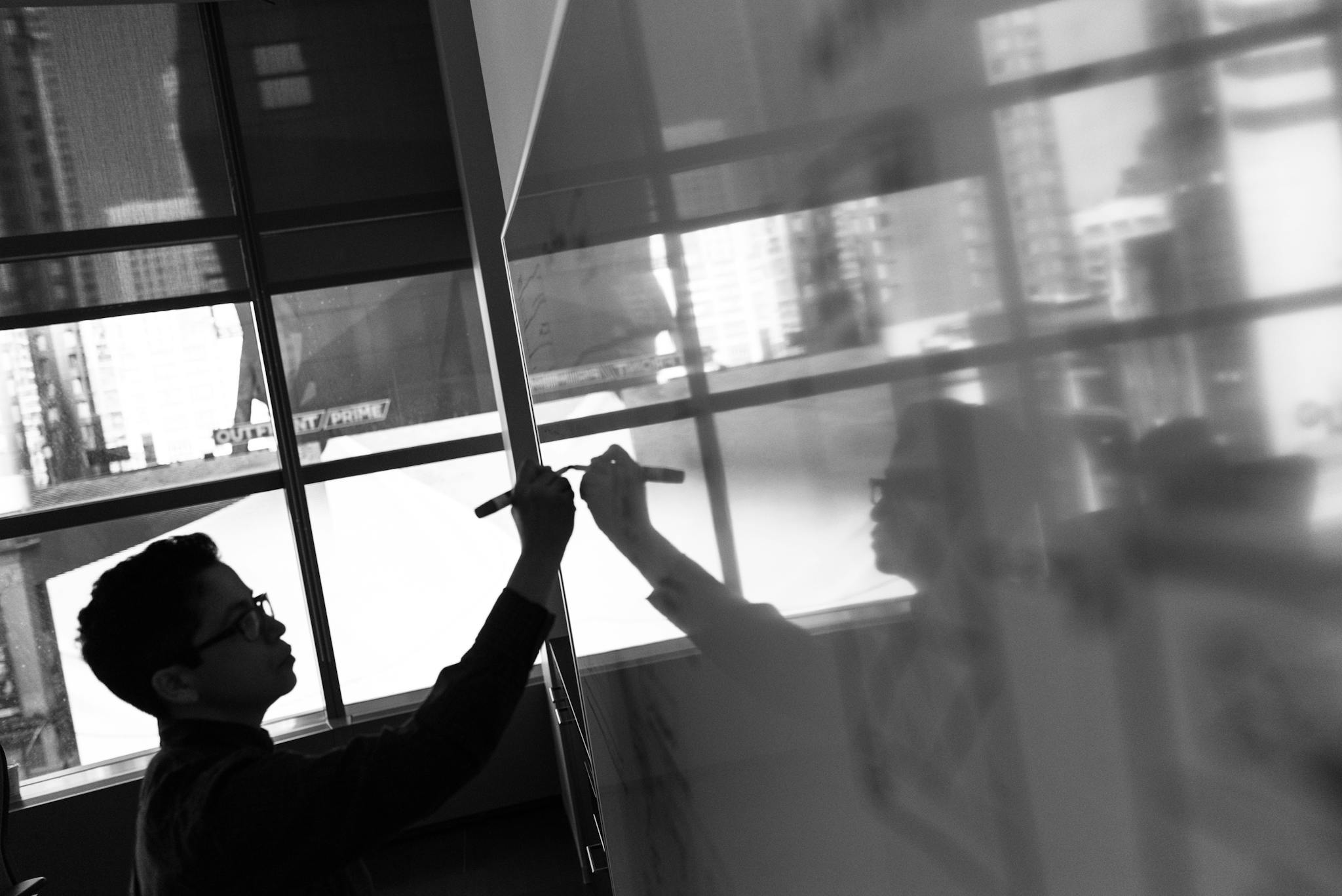 A person writing on a whiteboard with reflection in an office window.